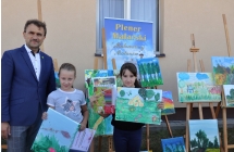 Dwie dziewczynki trzymają dyplomy. Obok nich mężczyzna w garniturze. Za nimi wystawa obrazów na sztalugach 