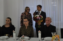 Mężczyzna mówiący do mikrofonu. Obok kobieta i inni goście siedzący na krzesełkach 